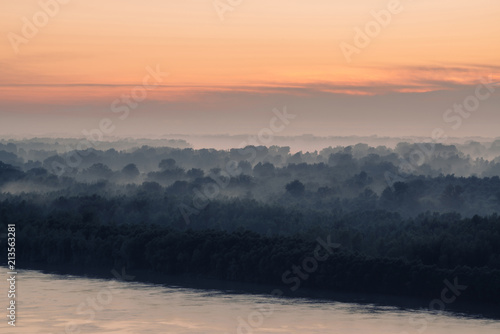 Mystical view on riverbank of large island with forest under haze at early morning. Eerie mist among layers from tree silhouettes. Morning atmospheric landscape of majestic nature in blue faded tones.