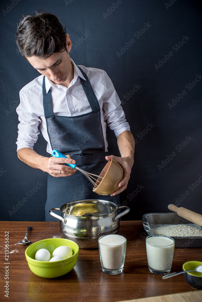 Young man chef in black apron cooking cake with ingredients on the table in the kitchen. Concept of men cook desserts Young man chef in black apron cooking cake with ingredients on the table in the kitchen. Concept of men cook desserts
