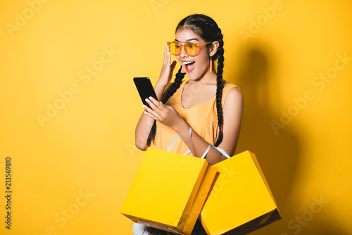 Beautiful young woman with shopping bags using her smart phone on yellow background.Shopaholic shopping Fashion.