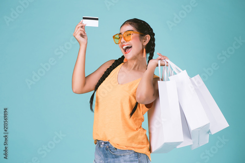 portrait of cheerful young brunette woman holding credit card and shopping bags over Blue background.