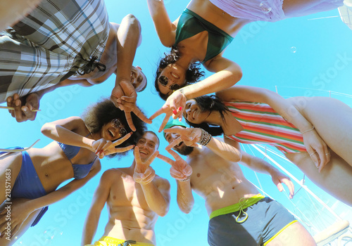 group of 6 young people gesturing looking down at camera gesturing peace with fingers. view from the bottom with blue sky background.