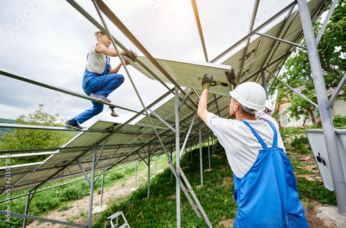 Installing of solar photo voltaic panel system. Three technicians lifting heavy solar module on high platform. Investment in alternative energy  money saving and professional construction concept.