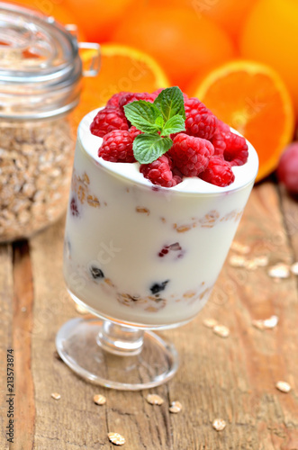 Greek yoghurt with oatmeal and fresh raspberries in a glass and mint leaves, full glass of oatmeals and oranges in background