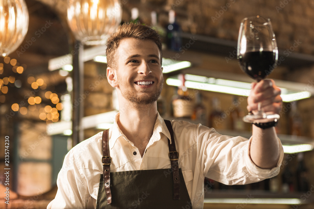 There you go. Upbeat young barman giving a glass of red wine to his customer and smiling at them brightly There you go. Upbeat young barman giving a glass of red wine to his customer and smiling at them brightly