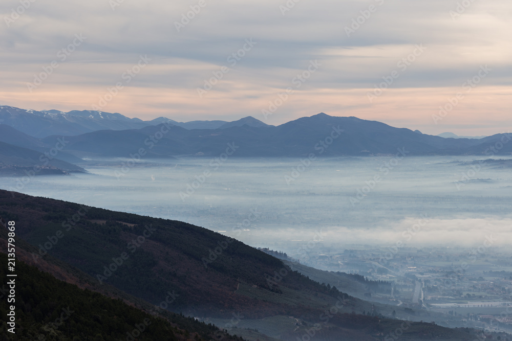 Beautiful aerial view of Umbria valley in a winter morning, with fog covering trees and house and warm colors in the sky