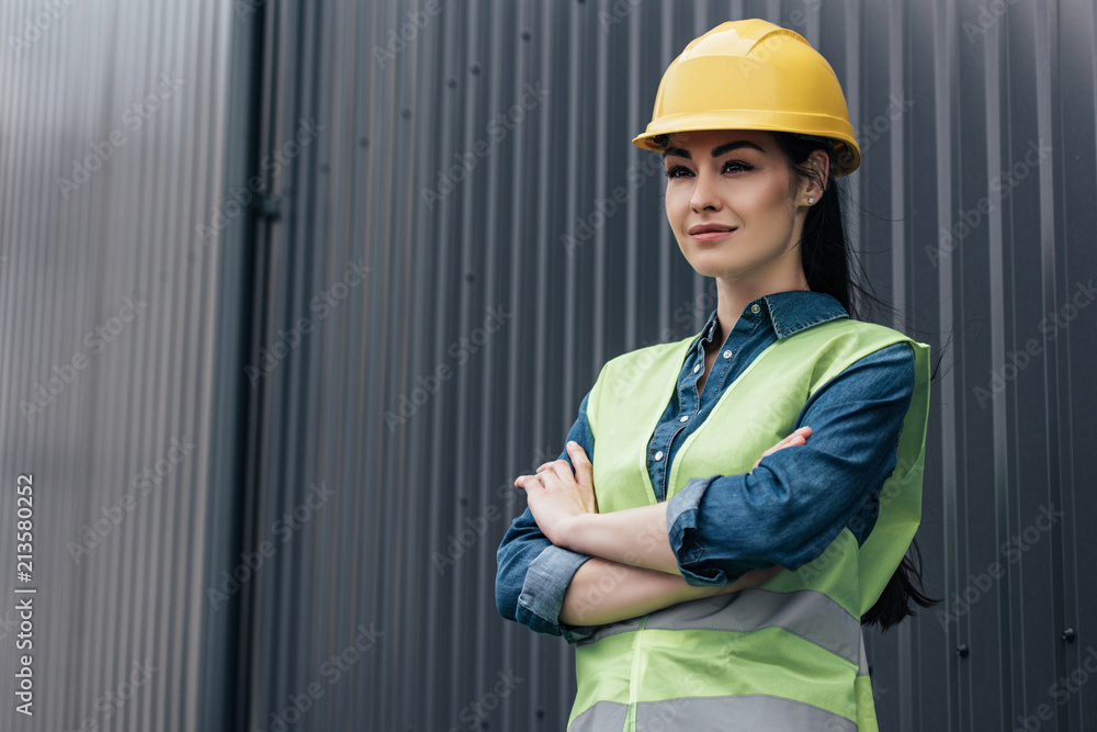 confident female architect in safety vest and helmet standing with crossed arms at wall confident female architect in safety vest and helmet standing with crossed arms at wall