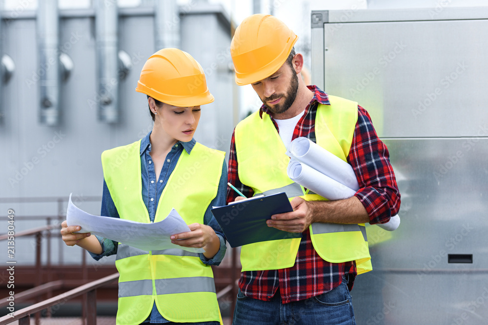 professional engineers in safety vests and hardhats working with blueprints and clipboard on roof professional engineers in safety vests and hardhats working with blueprints and clipboard on roof