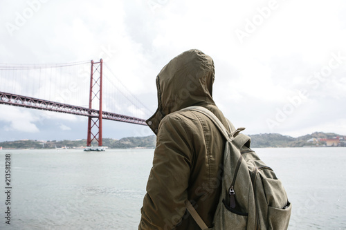 A young traveler or a tourist with a backpack on the waterfront in Lisbon in Portugal next to the 25th of April Bridge