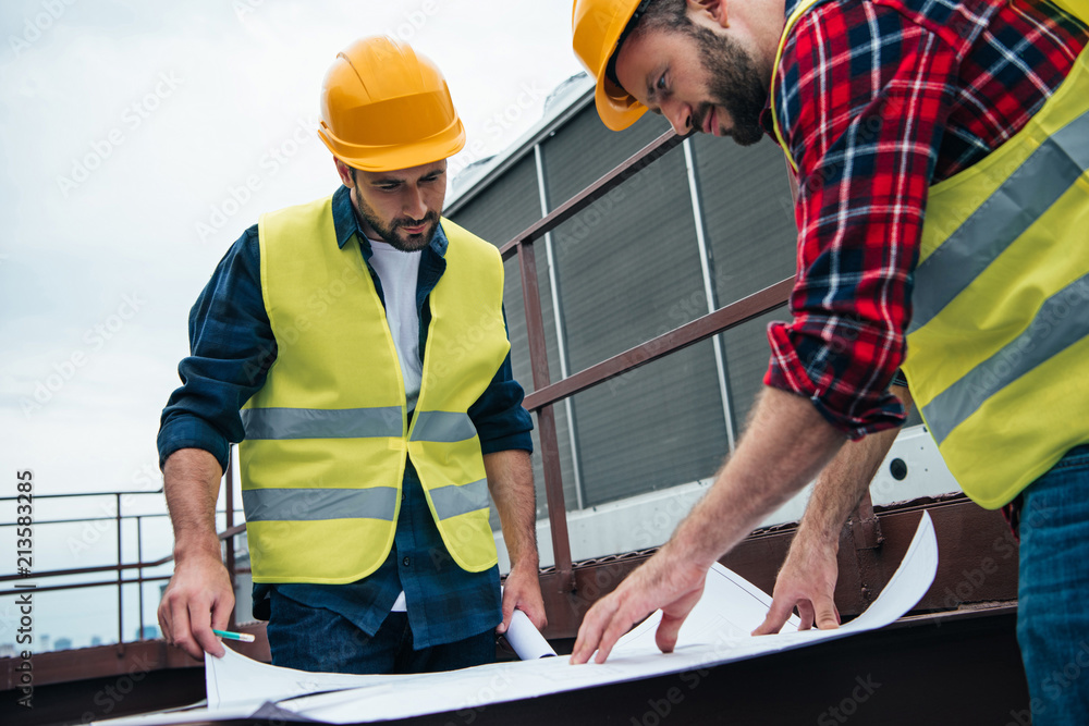 architects in safety vests and hardhats working with blueprints on roof architects in safety vests and hardhats working with blueprints on roof
