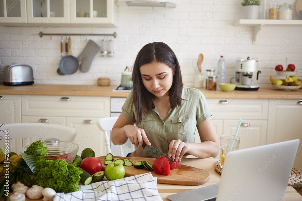 Cooking, culinary, quinine, food and nutrition concept. Indoor shot of hardworking young housewife with dark hair slicing red bell pepper and cucumber while making vegetable meal for dinner Cooking, culinary, quinine, food and nutrition concept. Indoor shot of hardworking young housewife with dark hair slicing red bell pepper and cucumber while making vegetable meal for dinner