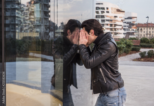Side view of man in leather jacket and jeans looking inside of window leaning to glass while standing on street 