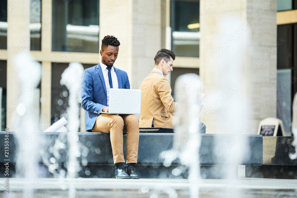 Serious concentrated handsome black male entrepreneur typing on laptop while examining data while sitting in city park outdoors