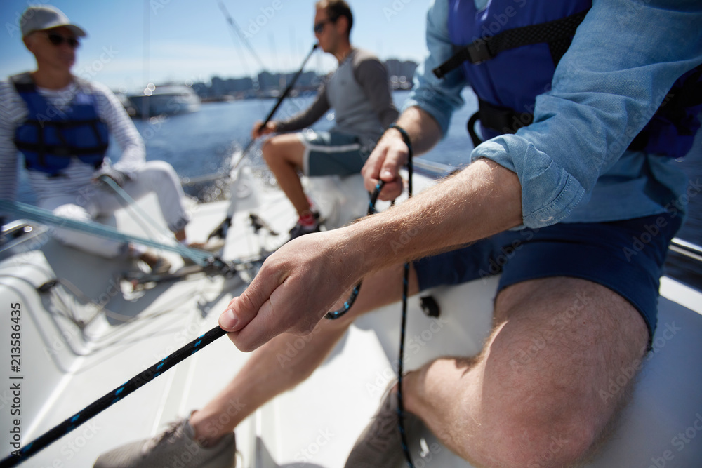 Fototapeta premium Close-up of male hands pulling rope of sailboat while mooring yacht and sitting with friends on deck