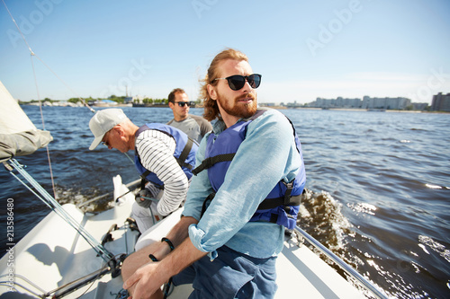 Content carefree young bearded man in sunglasses enjoying yachting with friends: he sitting on deck and contemplating seascape