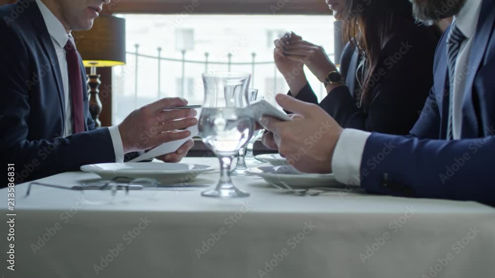Group of three business people sitting at table in cafe or restaurant and discussing documents when waiting for food during business lunch Group of three business people sitting at table in cafe or restaurant and discussing documents when waiting for food during business lunch