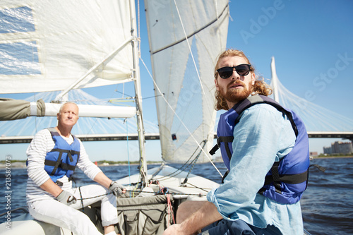 Serious handsome men in life jacket frowning from sun looking around while traveling by sail boat on river