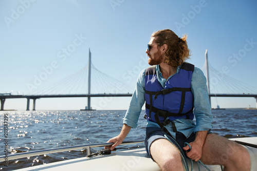Serious thoughtful handsome young guy with beard wearing sunglasses and life jacket admiring waterscape sitting on boat deck and holding railing