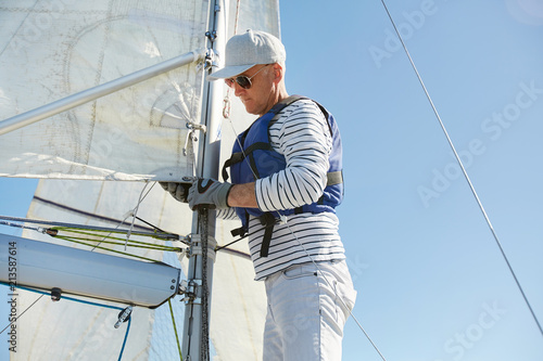 Serious skilled mature sailor in sunglasses and cap wearing stylish outfit fixing mainsail on mast with rope