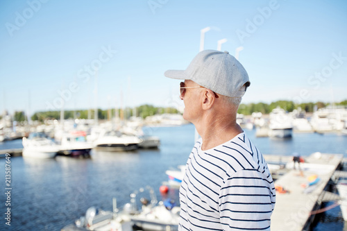 Pensive dreamy mature man in cap and sunglasses being on vacation looking at sailboats while standing on pier in yacht club © pressmaster