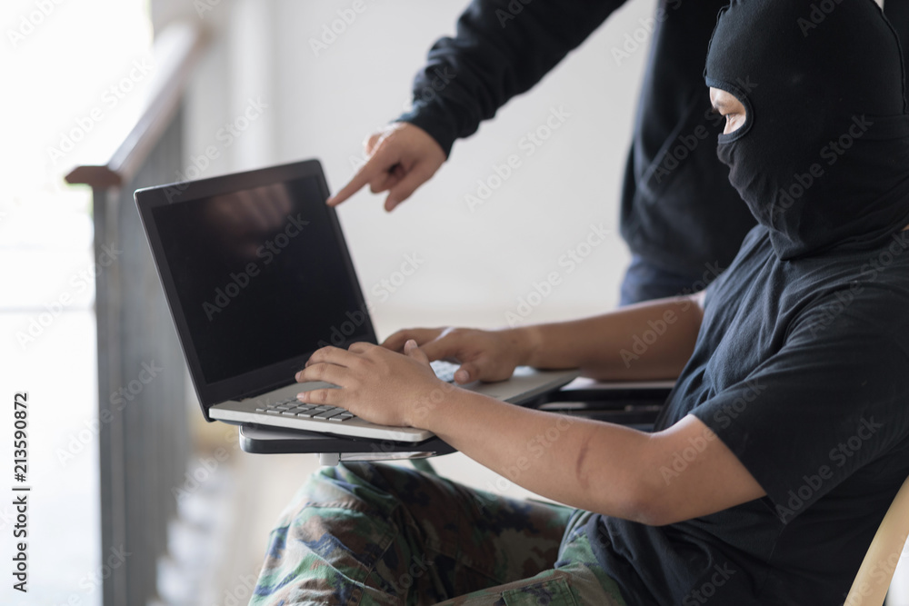 Two black masked hackers are using laptop. Hacker in the camouflage pant is sit with one's legs crossed and look at laptop. And the blue jeans hacker is standing and point at laptop. Selective focus a Two black masked hackers are using laptop. Hacker in the camouflage pant is sit with one's legs crossed and look at laptop. And the blue jeans hacker is standing and point at laptop. Selective focus a