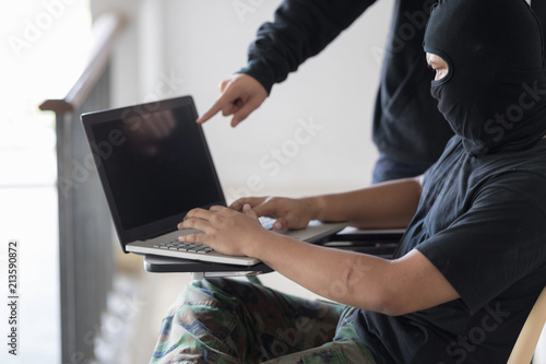 Two black masked hackers are using laptop. Hacker in the camouflage pant is sit with one's legs crossed and look at laptop. And the blue jeans hacker is standing and point at laptop. Selective focus a