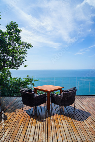 Table and rattan outdoor armchair setting on wooden deck and glass balcony with panoramic Andaman ocean sea view, green tree and blue sky background on sunny day