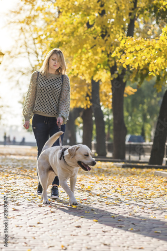A blonde is walking with a labrador in the park.