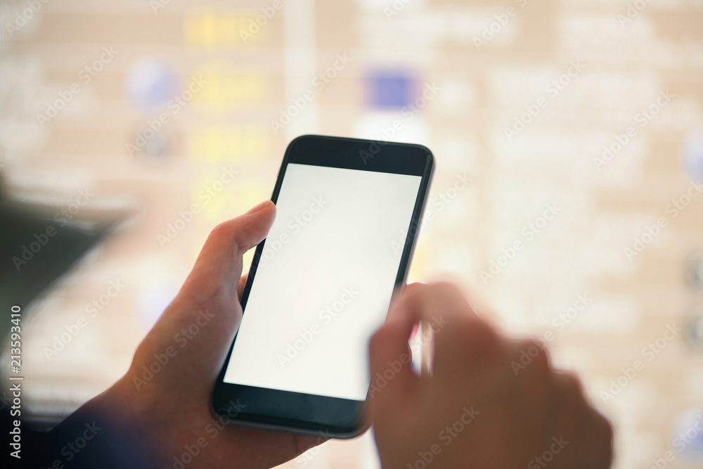 Mockup image of female hands holding black mobile phone with blank white screen over flight board in airport terminal