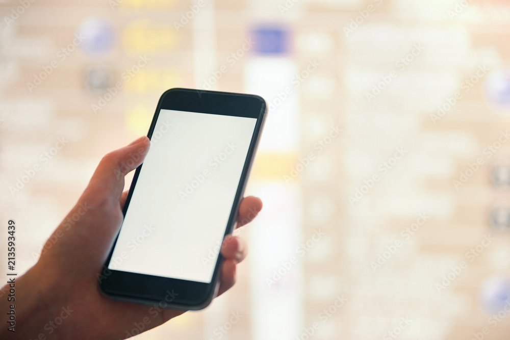 Mockup image of female hands holding black mobile phone with blank white screen over flight board in airport terminal