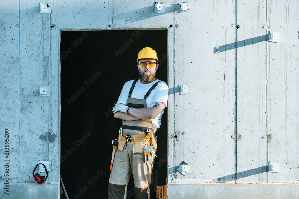 builder in protective googles and hardhat standing with crossed arms at construction site builder in protective googles and hardhat standing with crossed arms at construction site