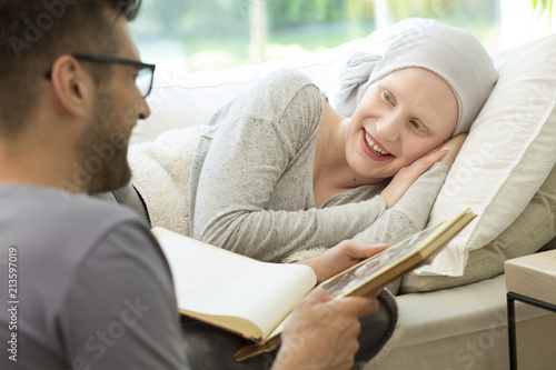 Husband reading book to smiling sick woman with headscarf photo