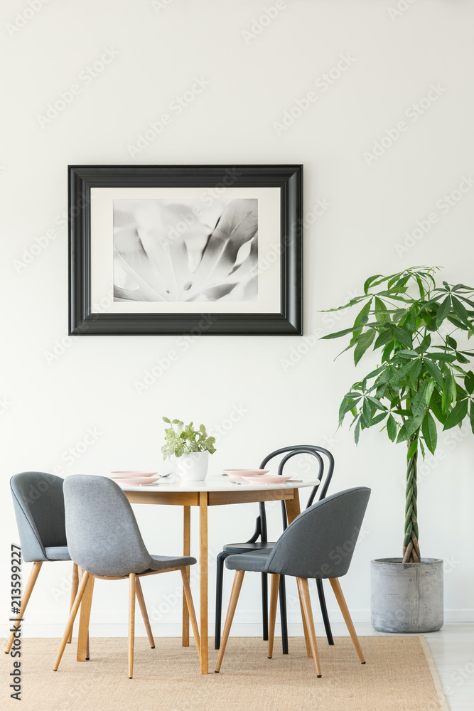 Real photo of a dining room interior with a table, chairs, tree and painting in a black frame