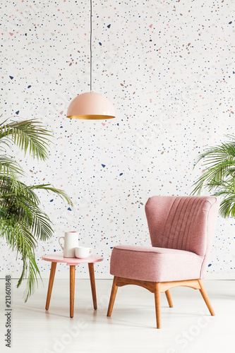 Pink lamp above table and armchair in patterned living room interior with plants. Real photo
