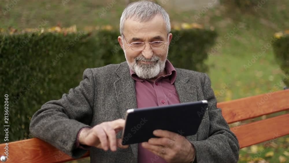 Grandpa use a tablet sitting in the park on the bench Grandpa use a tablet sitting in the park on the bench