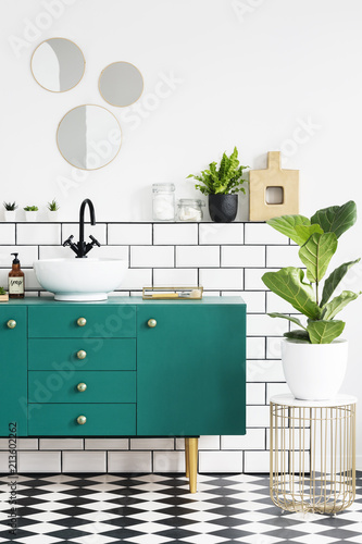 Green cabinet next to plant on gold table in modern bathroom interior with mirrors. Real photo
