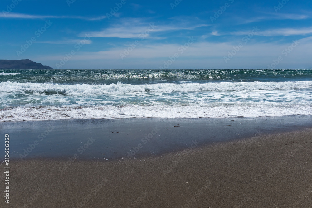 Background of coastal sand, sea waves against a blue sky with clouds and a mountain in the distance