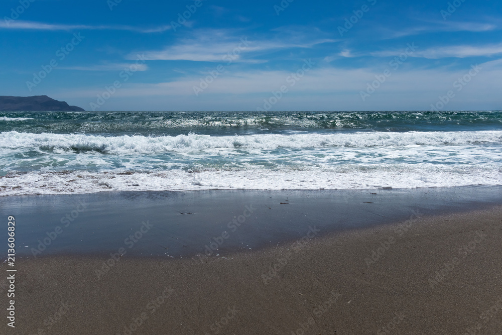 Background of coastal sand, sea waves against a blue sky with clouds and a mountain in the distance Background of coastal sand, sea waves against a blue sky with clouds and a mountain in the distance