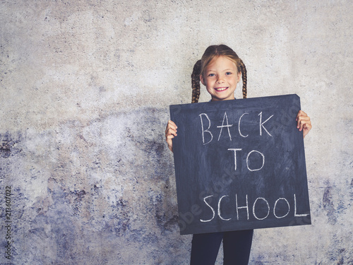 blond schoolgirl is holding blackboard with the words back to school in front of concrete background photo