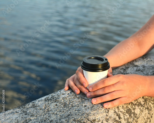 Hands of a girl holding a paper glass with tea or coffee while walking along the waterfront