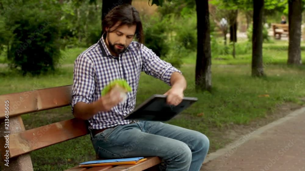 Man is walking to bench and sitting on it. He puts folders and burger on bench. Man puts laptop on legs and open it. Guy opens laptop, brushes hair and start to work. Then he takes burger and eat it. Man is walking to bench and sitting on it. He puts folders and burger on bench. Man puts laptop on legs and open it. Guy opens laptop, brushes hair and start to work. Then he takes burger and eat it.