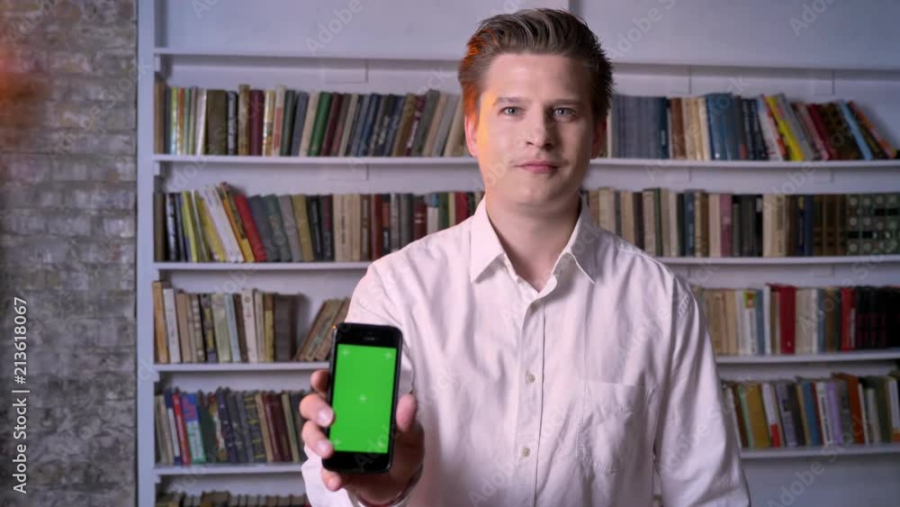 Young man is showing green screen of smartphone in library, watching at camera, communication concept Young man is showing green screen of smartphone in library, watching at camera, communication concept