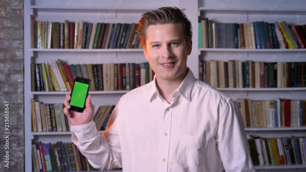 Young confident man is showing green screen of smartphone in library, watching at camera, communication concept Young confident man is showing green screen of smartphone in library, watching at camera, communication concept