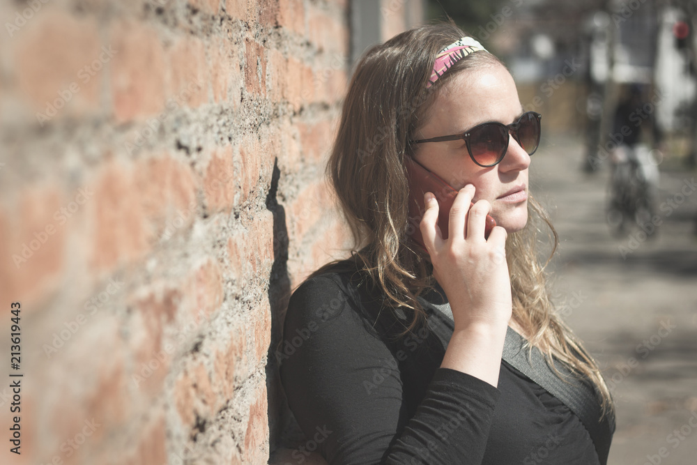 Serious young woman with smart phone on ear leaning on brown brick wall outdoor. Girl with sunglasses and cellphone on street. Vintage effect