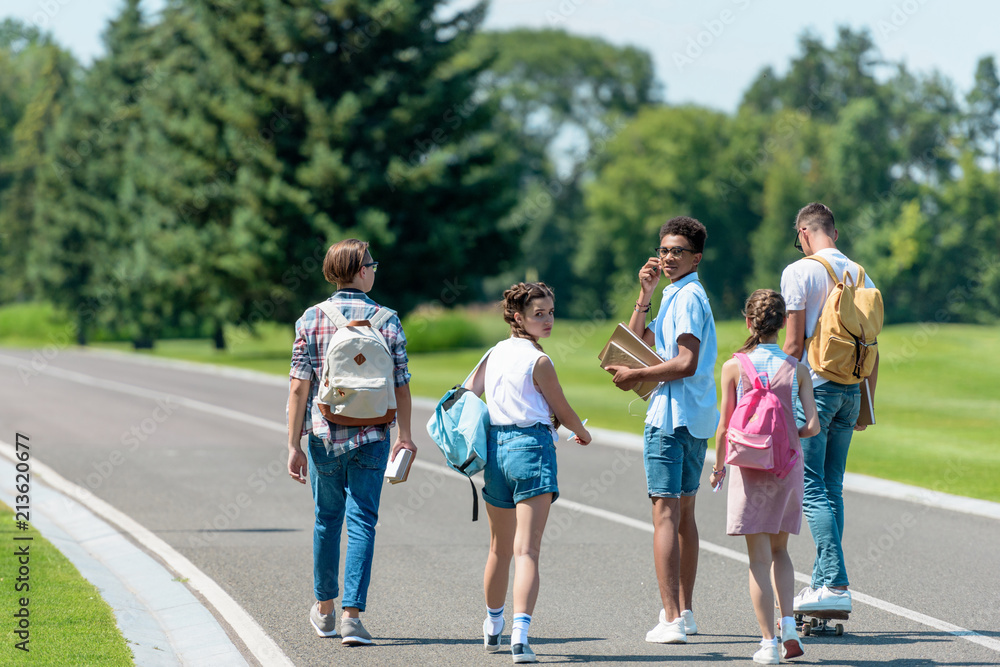 back view of multiethnic teenage friends with books and backpacks walking together in park back view of multiethnic teenage friends with books and backpacks walking together in park