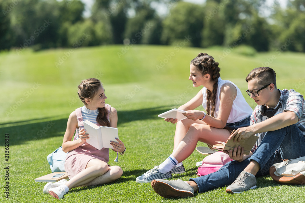 smiling teenage students with books and digital tablet sitting on grass and studying together in park