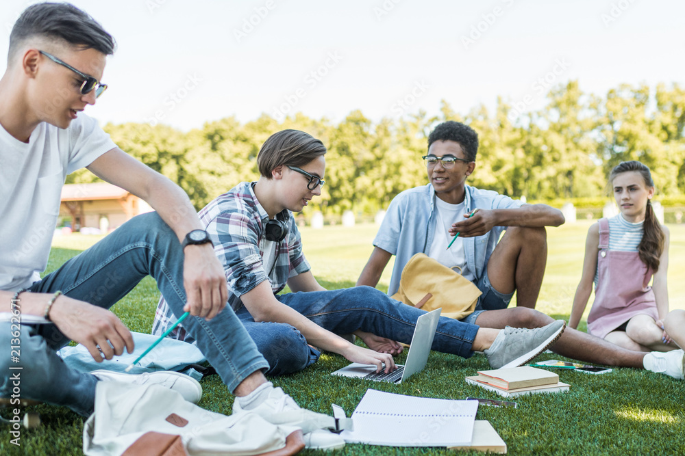 happy multiethnic teenagers sitting and talking while studying together in park happy multiethnic teenagers sitting and talking while studying together in park