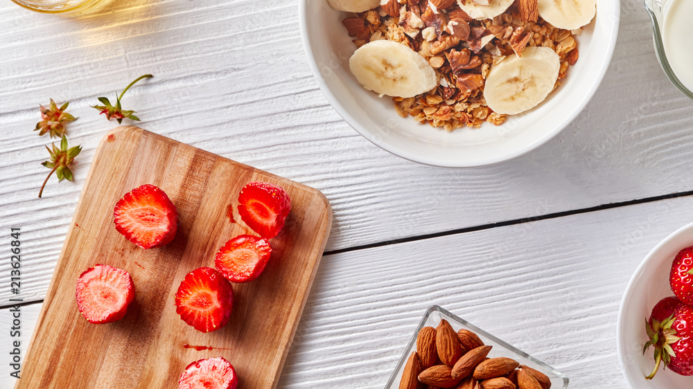 On the kitchen board halves of ripe strawberries, a plate with granola and banana, nuts of almonds on a white kitchen table. Vitamin breakfast On the kitchen board halves of ripe strawberries, a plate with granola and banana, nuts of almonds on a white kitchen table. Vitamin breakfast