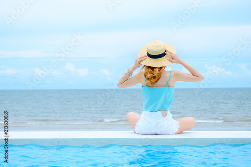 Young beautiful women wearing blue Spaghetti strap and bamboo hat sitting on edge of swimming pool enjoy life on tropical beach with blue sky.