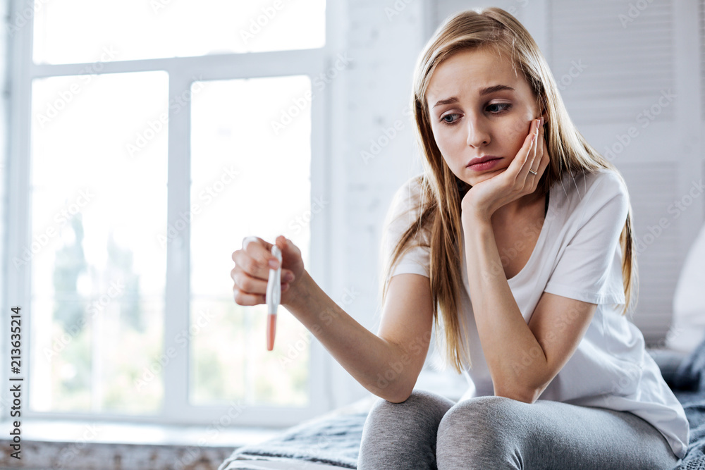 Bad mood. Unhappy young woman sitting and holding her pregnancy test