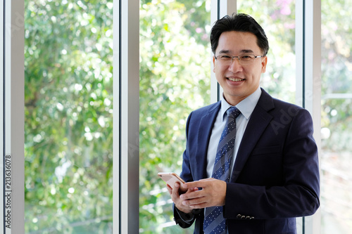 Asian Businessman holding smart phone with happy smiling face standing by windows, inside eco office building background, businessman on smart phone